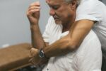 An older man sits while another man hugs his shoulder from behind in a comforting gesture with a watch visible on the arm of the hugger.
