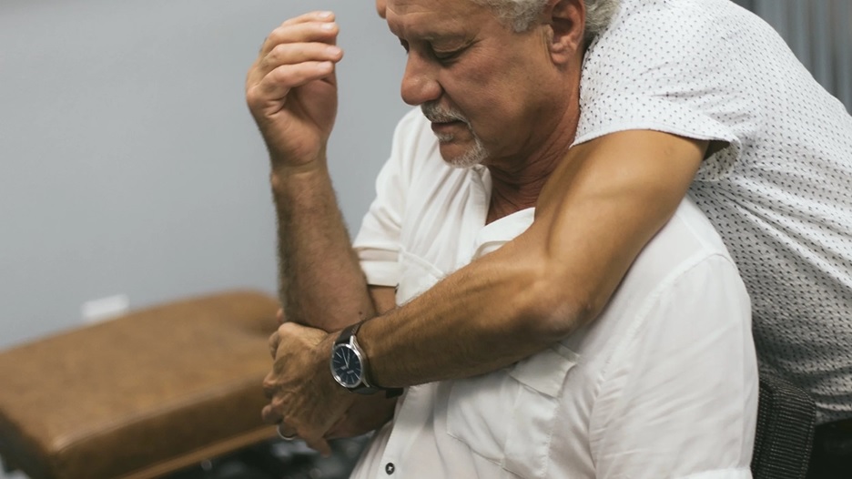 An older man sits while another man hugs his shoulder from behind in a comforting gesture with a watch visible on the arm of the hugger.